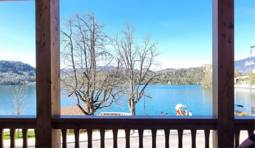 View of Lake Bled from Hotel Starkl, framed by a wooden balcony railing and support beams, with bare trees in the foreground and traditional wooden boats on the bright blue water. Hills and mountains form a scenic backdrop under a clear, sunny sky.