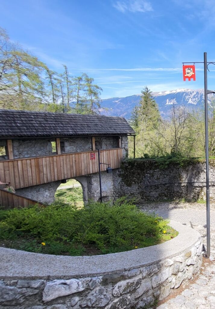 A rustic wooden bridge with stone supports leads through a forested area near a medieval structure, with snow-capped mountains rising in the background. A red castle-themed flag hangs from a nearby lamppost under a bright blue sky.