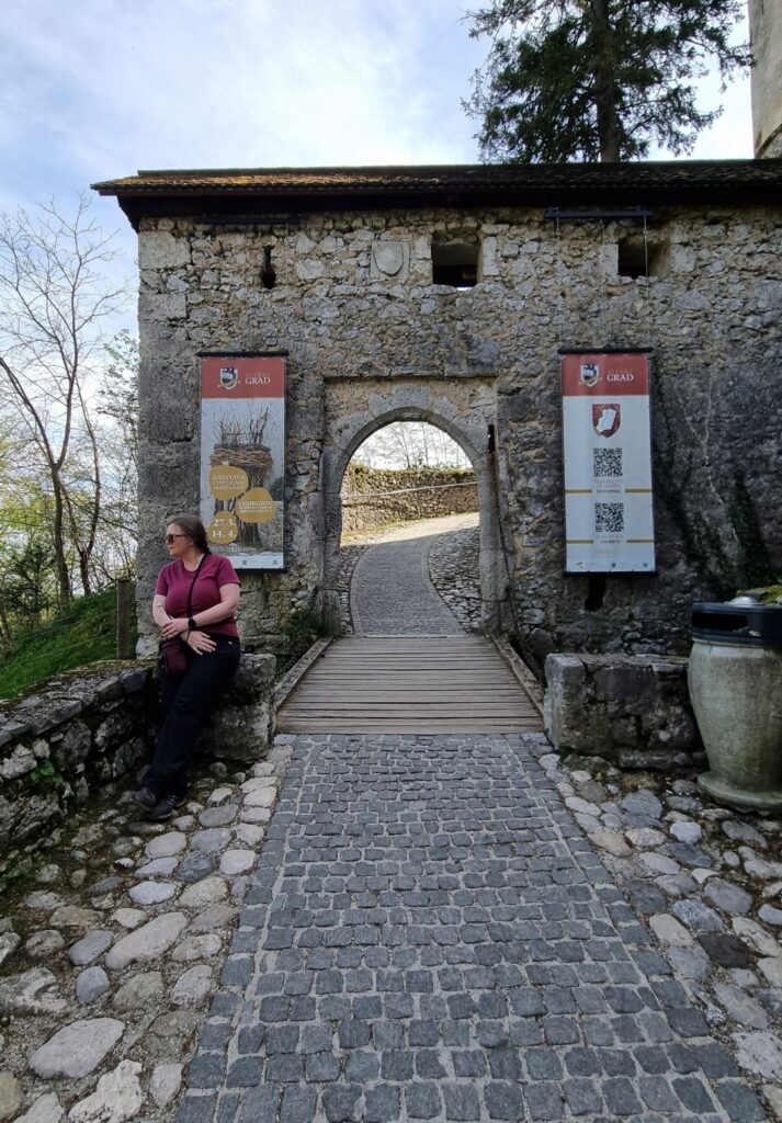 A woman leans casually against a stone wall outside the arched entrance of Bled Castle, flanked by informational banners. The cobblestone path and wooden ramp lead through the weathered stone gate into the historic site.