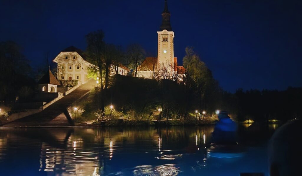 The Church of the Assumption on Bled Island glows warmly against the night sky, with lights illuminating the steep staircase and reflecting on the dark, still waters of Lake Bled.