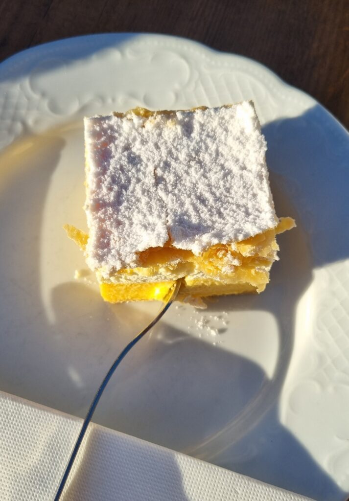 A square slice of traditional cream cake, known as kremšnita, sits on a white plate, topped with a thick dusting of powdered sugar and layered with golden puff pastry and custard. A fork is gently pressed into the cake, casting a shadow in the warm sunlight.