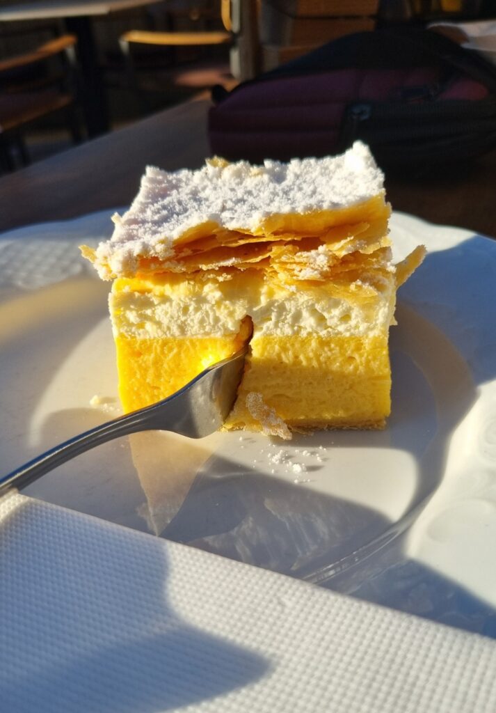 A close-up side view of a slice of kremšnita, showcasing its airy whipped cream, rich yellow custard, and crisp layers of puff pastry, all topped with powdered sugar. The fork has cut into the side, revealing the soft interior textures against a white plate.