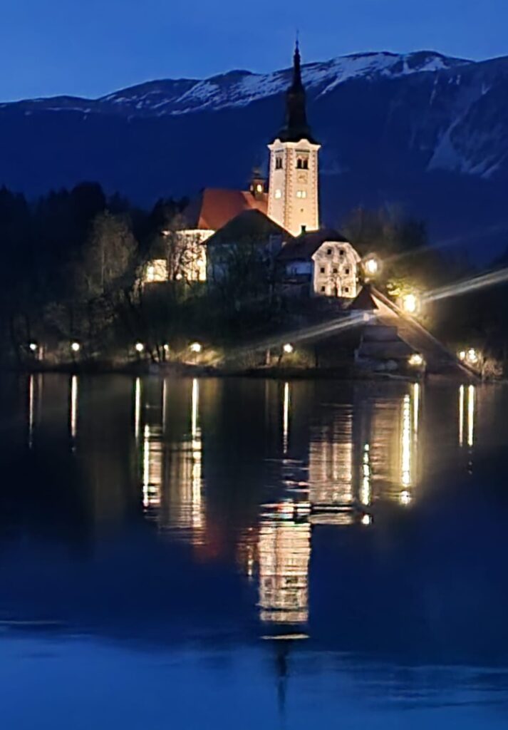 A close-up night view of the Church of the Assumption on Bled Island, with its tower and surrounding buildings lit up and casting shimmering reflections on the lake below.
