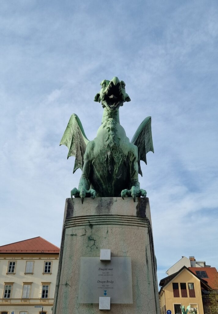 A close-up shot of the iconic green copper dragon statue on Ljubljana’s Dragon Bridge, perched on a stone pedestal under a partly cloudy sky.
