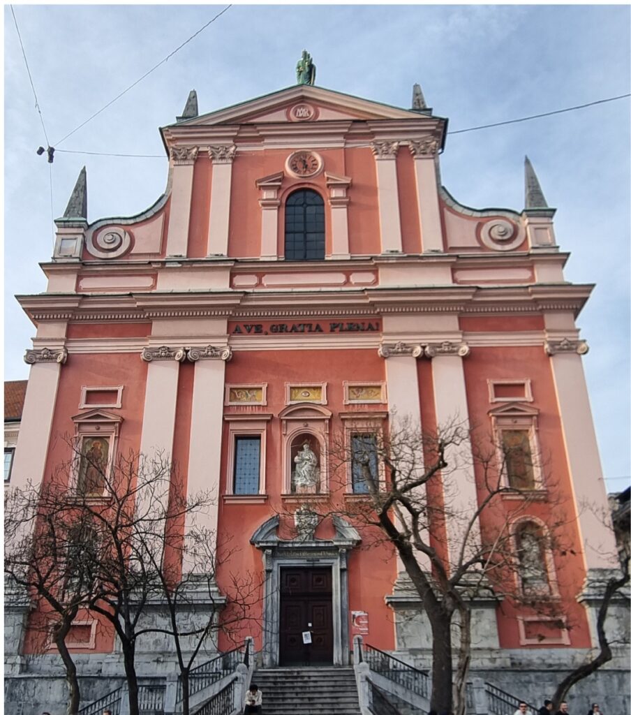 The vibrant pink-and-white Baroque facade of the Franciscan Church of the Annunciation in Ljubljana, with bare trees lining the entrance stairs.