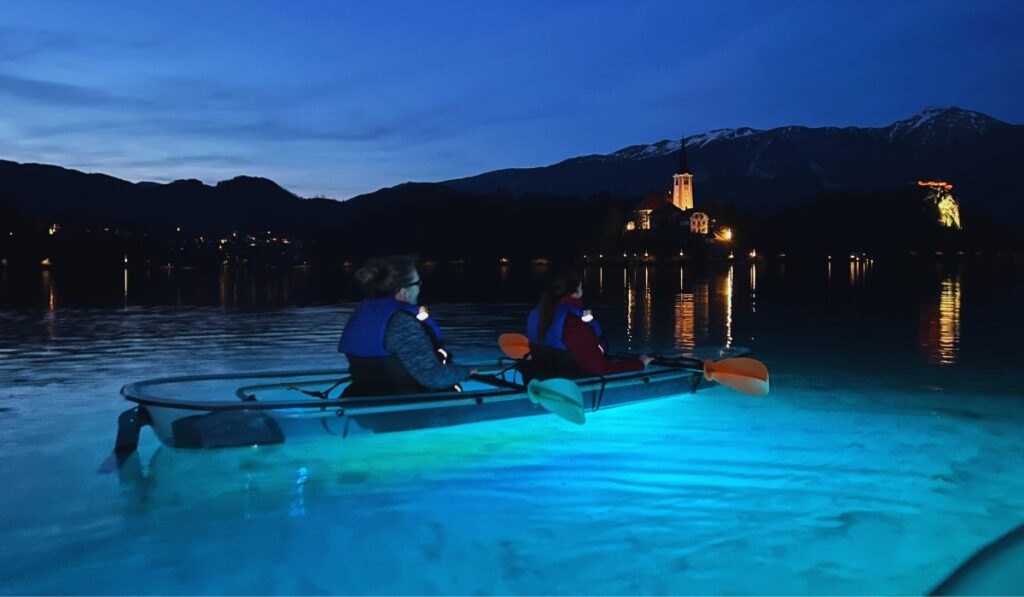 Sitting in a clear-bottom kayak glowing with blue LED lights, paddling across Lake Bled at twilight with views of the illuminated island church and Bled Castle in the distance.