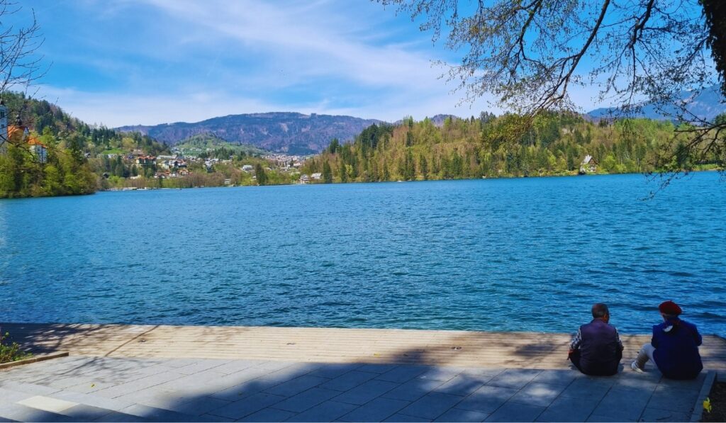 Two people sit on a shaded lakeside promenade overlooking the vivid blue waters of Lake Bled, with forested hills and scattered homes across the water. The serene setting is framed by overhanging tree branches and distant mountains under a sunny sky.