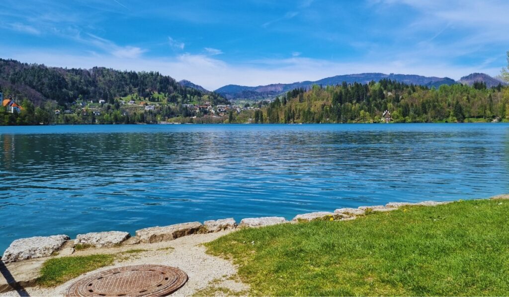 A peaceful lakeside scene at Lake Bled with crystal-clear turquoise water, green hills dotted with homes, and a stone path lined with grass and a manhole cover in the foreground. Rolling mountains frame the vibrant spring landscape under a bright blue sky.