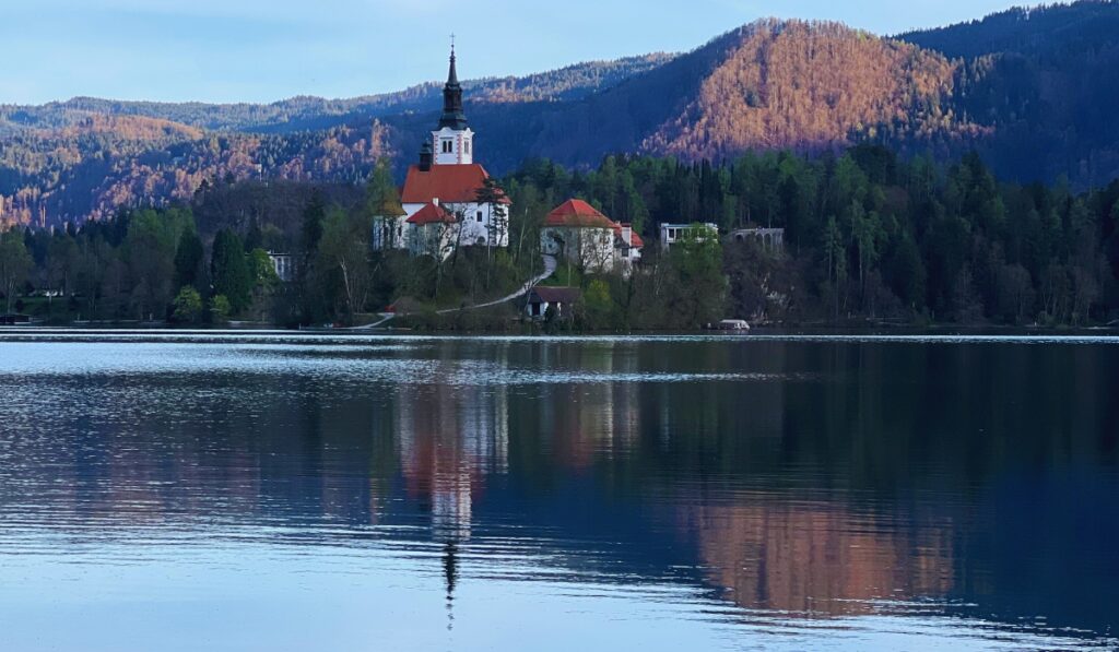 Lake Bled church with a tall spire and red roof stands on the shore of Lake Bled, surrounded by dense forest and reflected perfectly in the still water. Warm sunlight casts a gentle glow on the distant mountains, enhancing the serene atmosphere.