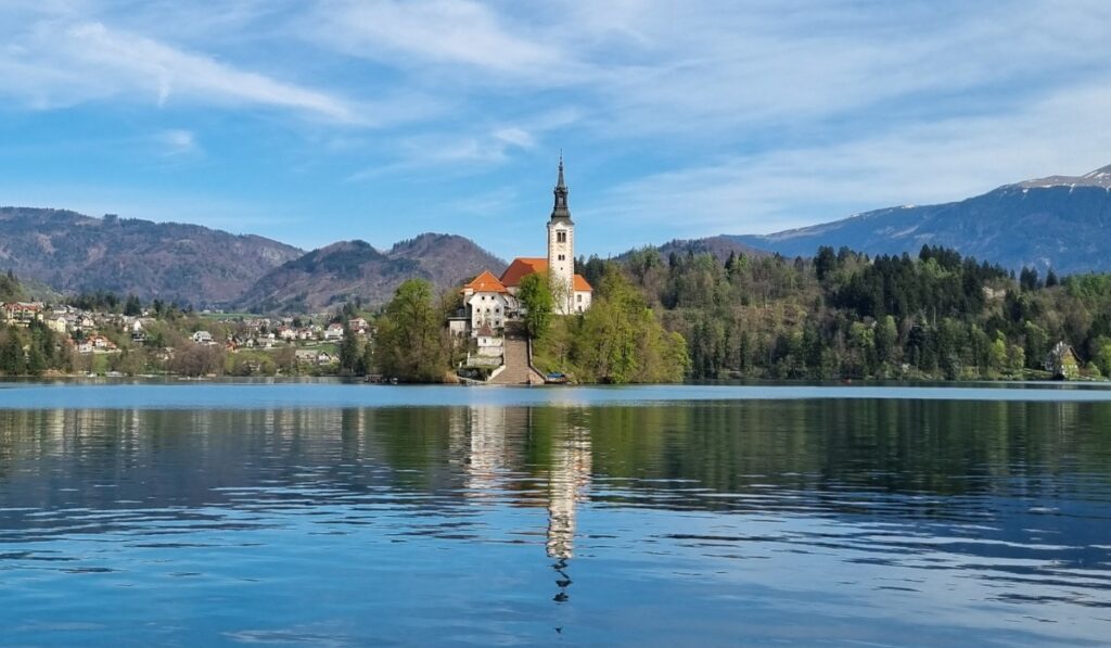 The Church of the Assumption stands tall on Bled Island, its white tower and red roof mirrored in the calm waters of Lake Bled. Surrounding hills and scattered homes create a picturesque backdrop beneath a partly cloudy spring sky.