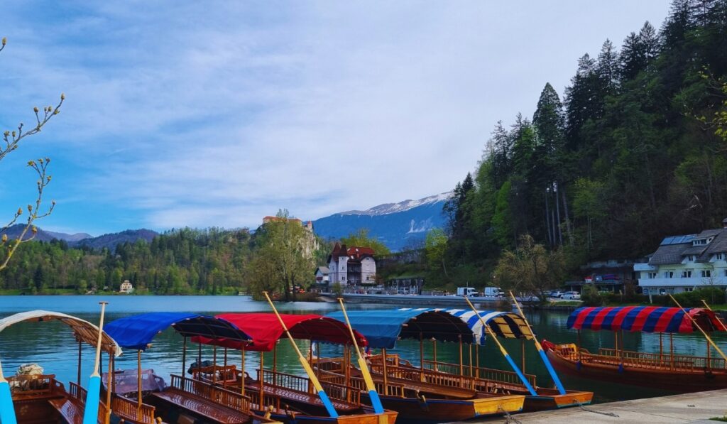 Traditional wooden boats with colorful canopies are docked on the calm waters of Lake Bled, with forested hills, a quaint lakeside village, and snow-capped mountains in the background. The clear blue sky and still lake reflect a peaceful alpine setting.
