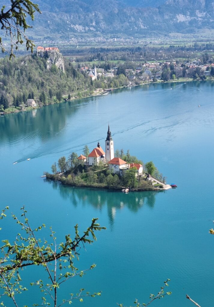 Aerial view of Bled Island in the middle of Lake Bled, featuring the Church of the Assumption with its iconic bell tower surrounded by turquoise water. In the distance, Bled Castle sits atop a cliff with the Julian Alps and town of Bled in the background.