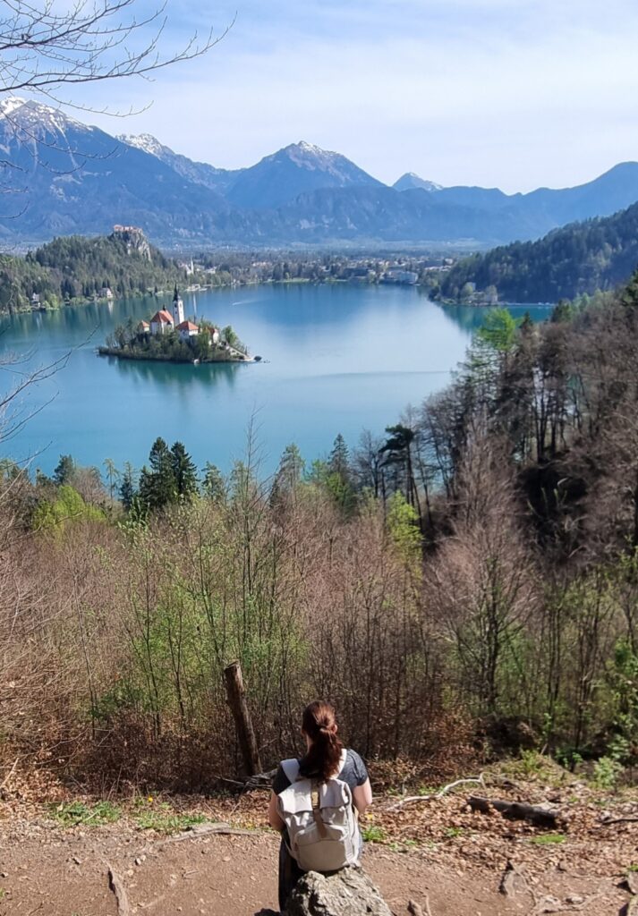 Me sitting on a rock overlooking Lake Bled and its small island with the church, framed by distant mountains and early spring trees. The elevated viewpoint captures a panoramic, peaceful vista.
