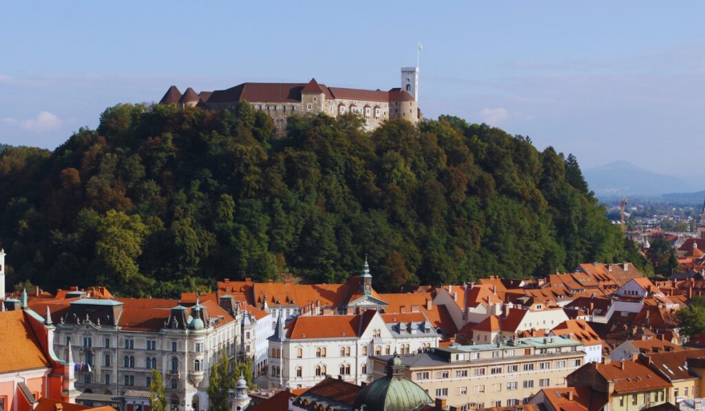 A panoramic view of Ljubljana Castle perched atop a lush green hill, overlooking the old town's red-roofed buildings and elegant historic architecture. The white watchtower flies the Slovenian flag under a clear blue sky.