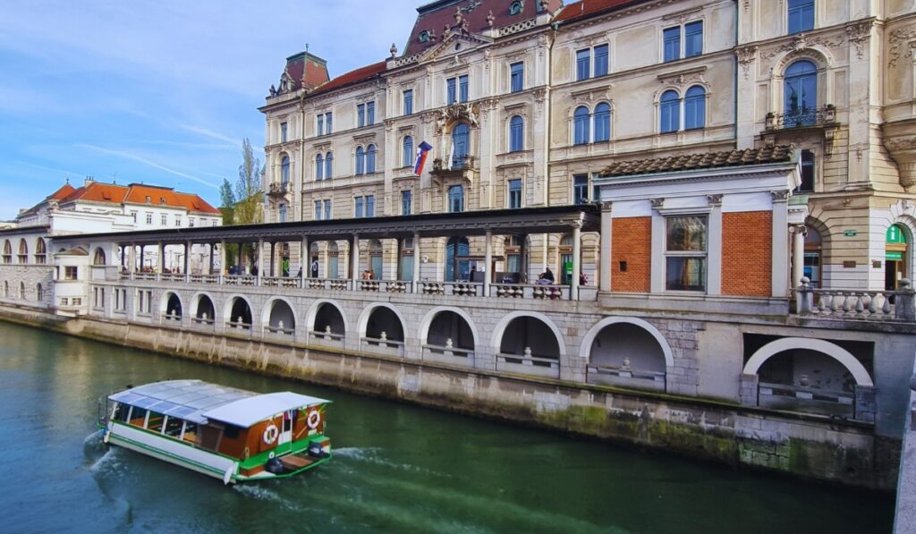 A green-and-white riverboat cruises down the Ljubljanica River beside the Central Market Colonnade, with an elegant historic building flying the Slovenian flag.