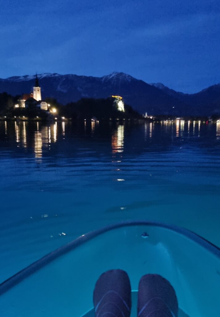 A view from inside a glowing kayak on Lake Bled at dusk, with feet visible in the foreground and the lit-up island church and Bled Castle reflecting on the calm water ahead.