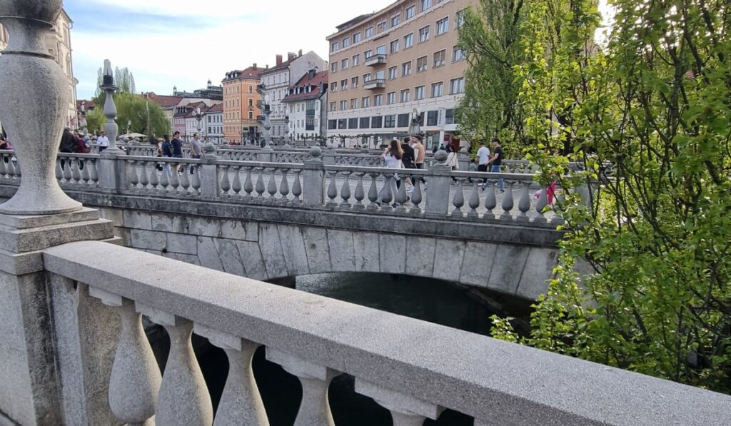 A wide view of Ljubljana’s Triple Bridge filled with pedestrians, framed by classic European buildings and lush green trees along the river.