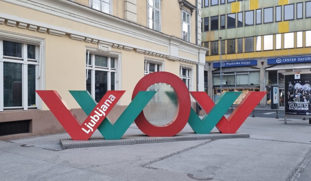A large colorful “WOW” sign in red and green with the word “Ljubljana” written on one of the letters, placed on a city sidewalk near historic and modern buildings.