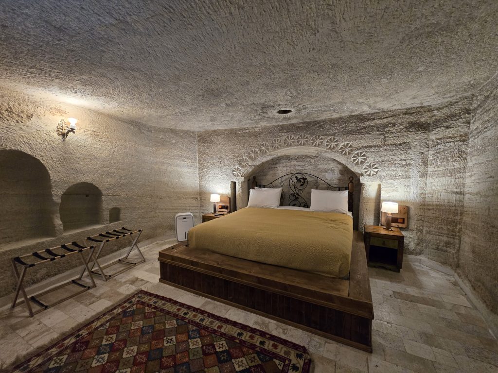 Cozy stone-carved cave hotel room with a low wooden platform bed covered in a mustard-yellow duvet, flanked by bedside tables and warm lighting. The textured walls and arched stonework behind the headboard create a rustic and authentic Cappadocian cave stay atmosphere.