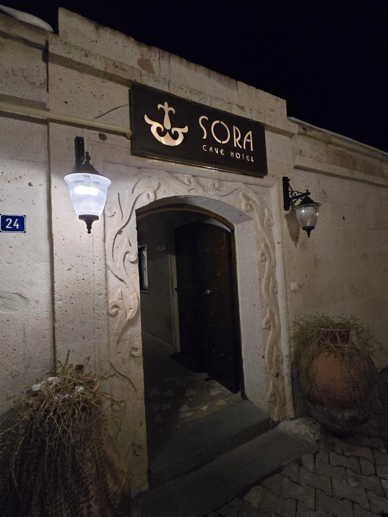 Nighttime view of the entrance to Sora Cave Hotel in Cappadocia, featuring a warmly lit stone archway with decorative carvings and a wooden sign above the door. Two wall lanterns and ceramic pots with dried plants frame the doorway.