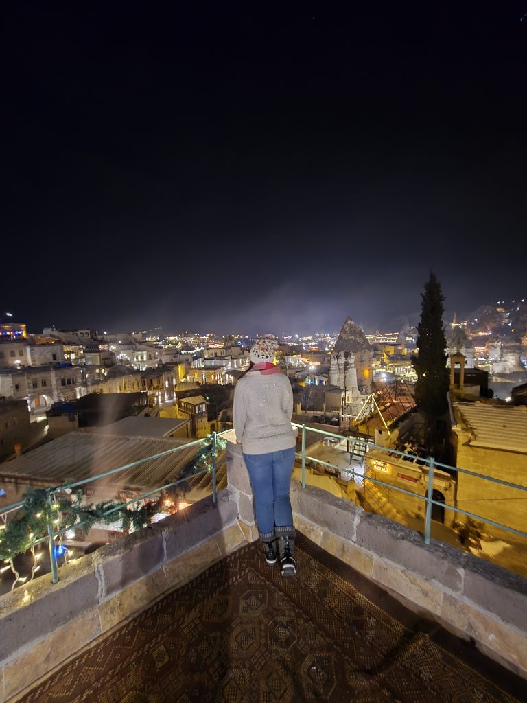 Me wearing a winter hat, scarf, and boots stands on a rooftop terrace overlooking a warmly lit cityscape at night in Cappadocia, Turkey. Fairy chimneys and stone buildings glow under ambient lights, with a patterned rug underfoot and festive garland on the railing.