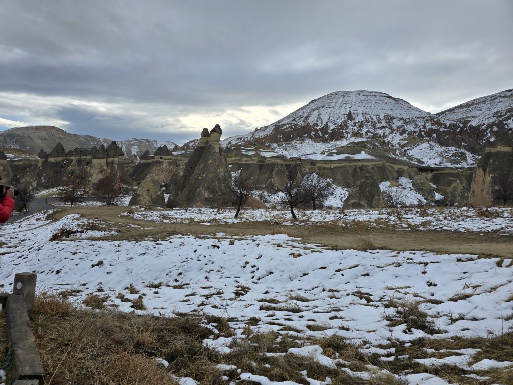 Snow-dusted fairy chimneys rise from a rugged landscape beneath a cloudy winter sky in Cappadocia, Turkey. A person in a red coat captures the scenery with a camera from the roadside.