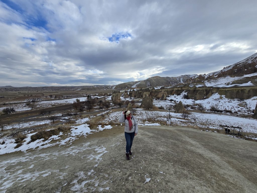 Me in a pink scarf and white jacket stands on a snowy overlook, gazing out across the expansive, snow-speckled rock formations and valley of Cappadocia in winter. The cloudy sky and rolling terrain emphasize the region's natural beauty.