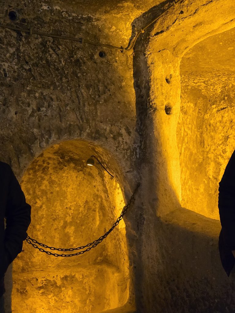 Dimly lit stone chamber in Cappadocia's underground city with a small, rounded alcove blocked off by a black chain. Warm yellow lighting highlights the rough texture of the carved rock walls.