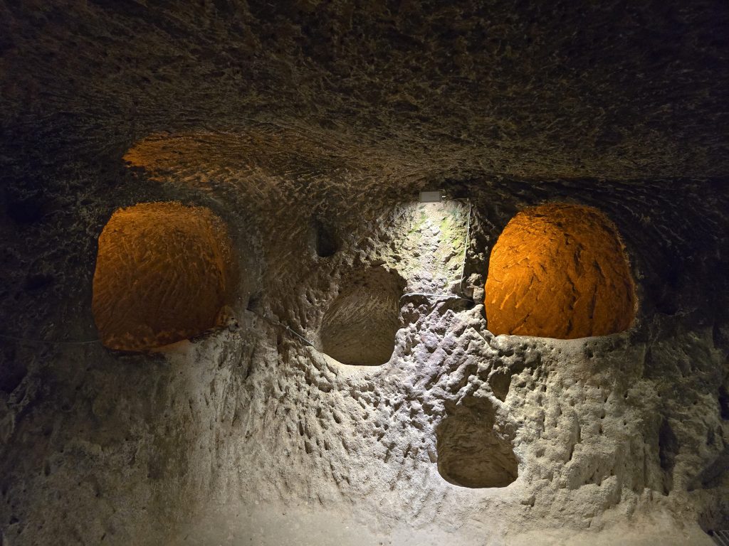A room carved from volcanic stone featuring three arched openings with orange lighting and several smaller holes below, part of an ancient underground city in Cappadocia. The space shows signs of centuries-old excavation and wear.