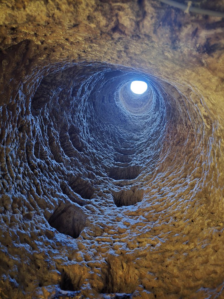 View from the bottom of a narrow vertical tunnel carved into rock, with a circular opening to daylight at the top and carved footholds spiraling upward. Likely used as a ventilation shaft or emergency escape route in Cappadocia’s underground cities.