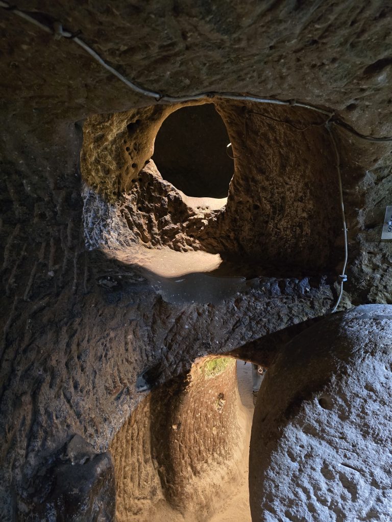 Rock-cut tunnel in an underground city showing a circular doorway and a partially visible millstone-style rolling door. Carved pathways and worn surfaces highlight the city’s complex subterranean architecture.