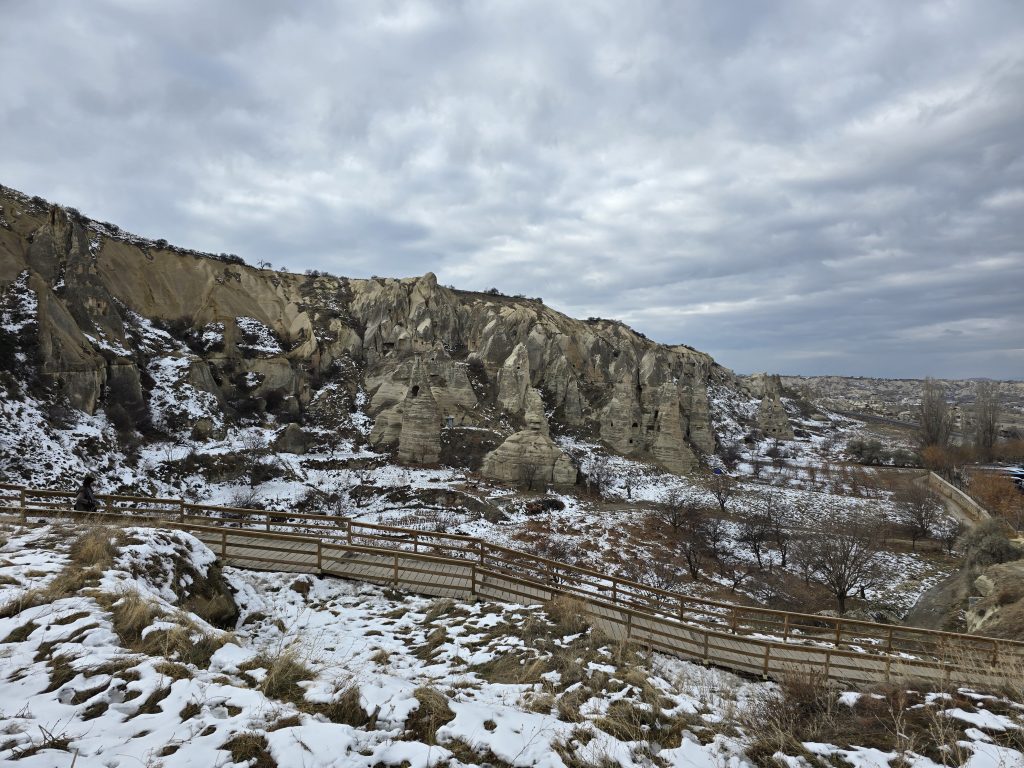 Snow-dusted valley surrounded by steep rock formations in Cappadocia, with a wooden boardwalk winding through the terrain. A cloudy winter sky looms above the dramatic landscape, while a lone visitor walks along the path.