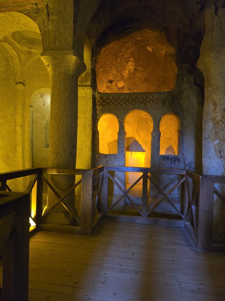 Warmly lit interior of a cave church in the Göreme Open-Air Museum, featuring arched stone architecture and a wooden viewing platform. The walls show faint carvings and traces of ancient frescoes.