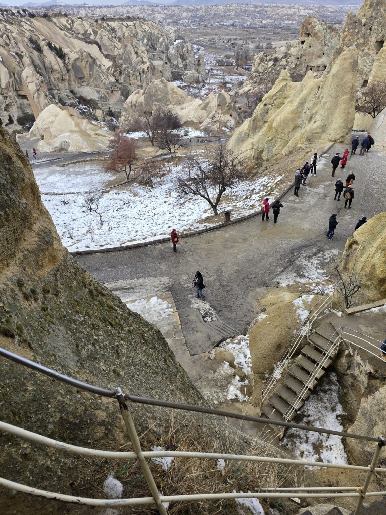 Looking down from a staircase carved into the rock at the Göreme Open-Air Museum, visitors walk along a snowy path between dramatic cliffside formations. The vast valley and other fairy chimneys are visible in the background.