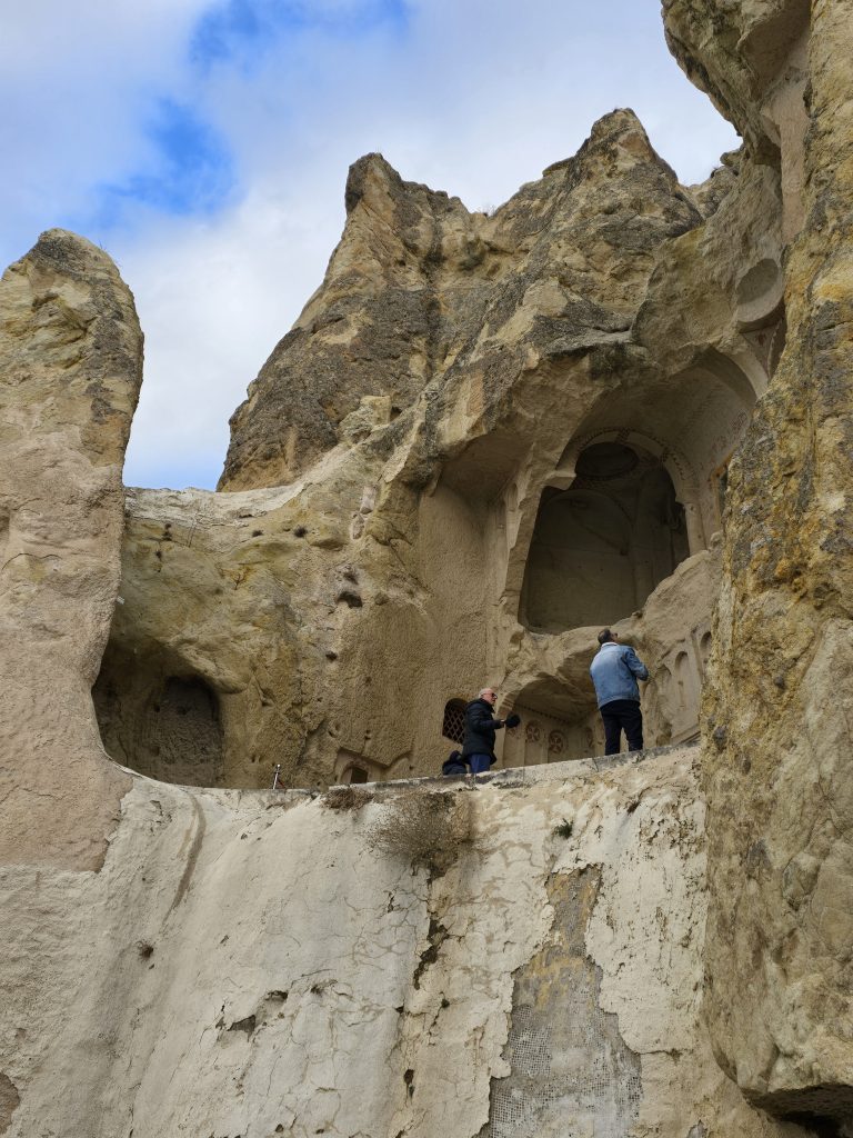 Two visitors stand on a high ledge examining the carved facade of a rock-hewn church at the Göreme Open-Air Museum. The structure features arched recesses and intricate details set into the rugged cliffside.