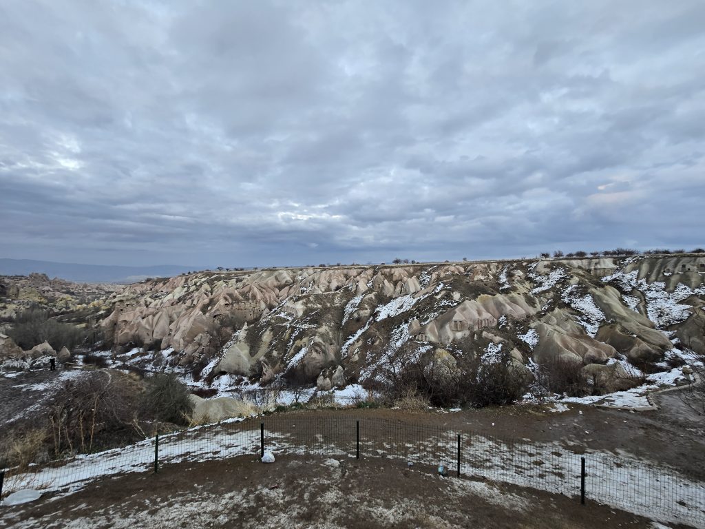A dramatic winter landscape in Cappadocia, Turkey, shows snow-dusted valleys and soft pink rock formations beneath a cloudy sky. A fence runs along the foreground, separating the viewer from the undulating terrain of eroded tuff hills.