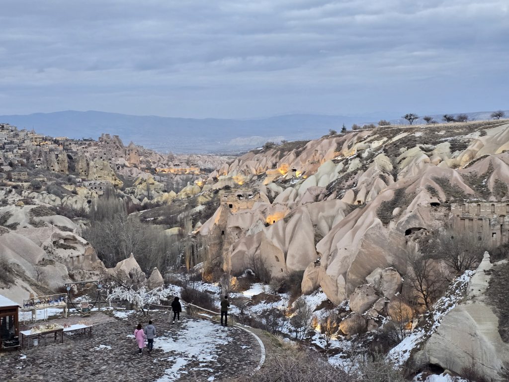 Overlooking Cappadocia’s illuminated cave dwellings and fairy chimneys at dusk, with families walking on a snow-covered stone terrace. The softly lit rock formations contrast against the moody sky and distant mountains, creating a magical winter evening scene.