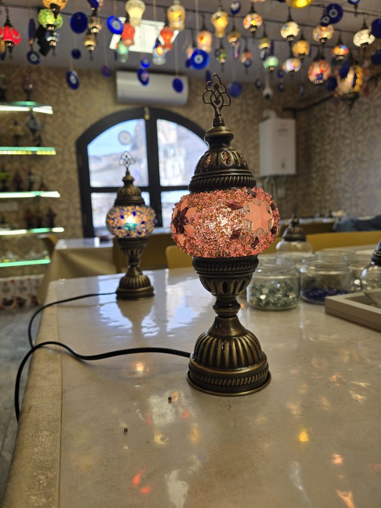A table lined with handmade mosaic lamps featuring intricate pink and blue glasswork, set in a cozy workshop with hanging Turkish lamps and evil eye charms suspended from the ceiling. The space glows warmly with ambient light and decorative patterns.