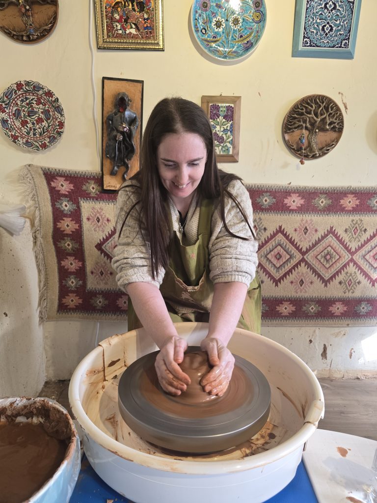 A woman smiling as she shapes clay on a pottery wheel in a cozy studio adorned with colorful ceramic art and traditional Turkish decor. Her hands are wet with clay, focused on forming the base of a pot.