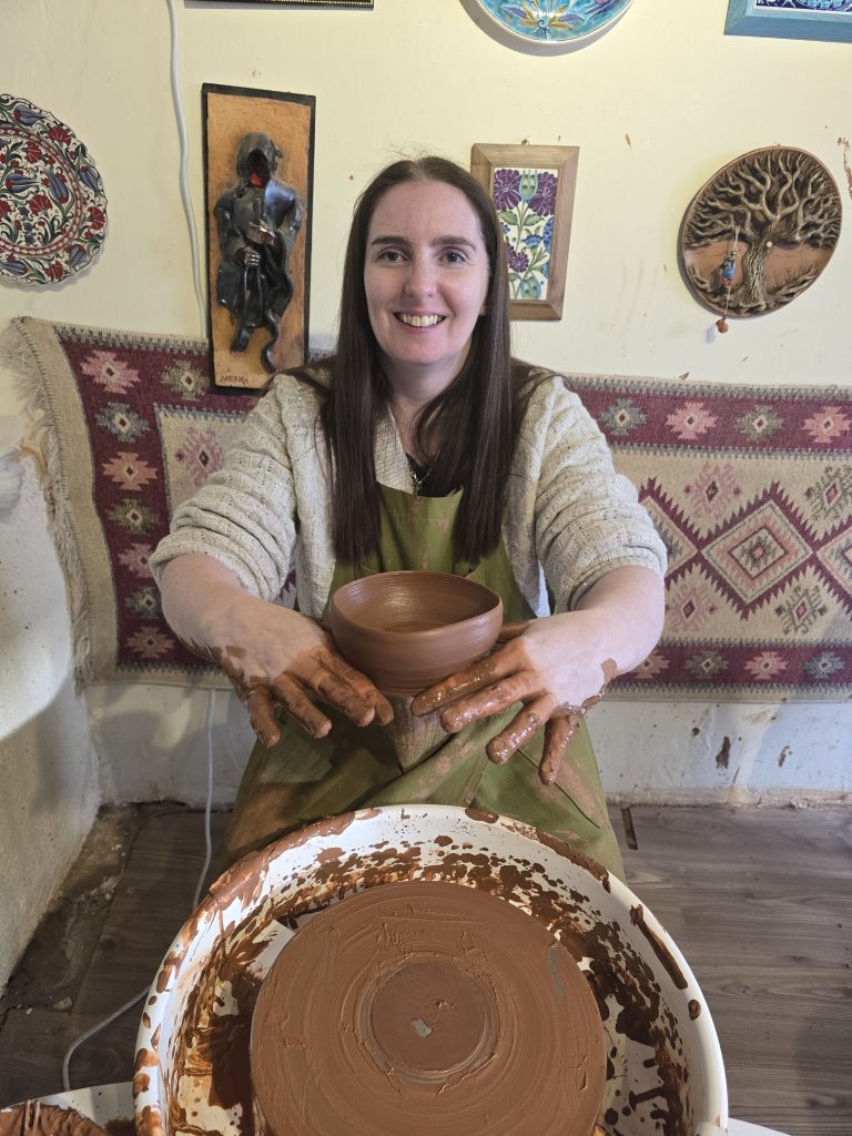 The same woman proudly holds a freshly made clay bowl, smiling at the camera. Her hands are still covered in clay, with the pottery wheel and a finished circular imprint of the bowl’s base in front of her.