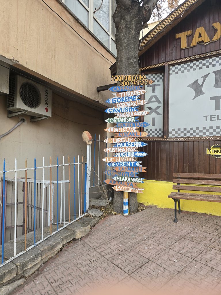 A colorful wooden signpost mounted on a tree points in various directions, listing popular destinations in Cappadocia, Turkey such as Göreme, Ürgüp, Kaymaklı, and Ihlara Vadisi. The sign is next to a bench and a “Yurt Taxi” office with a rustic exterior and a sidewalk made of red brick.