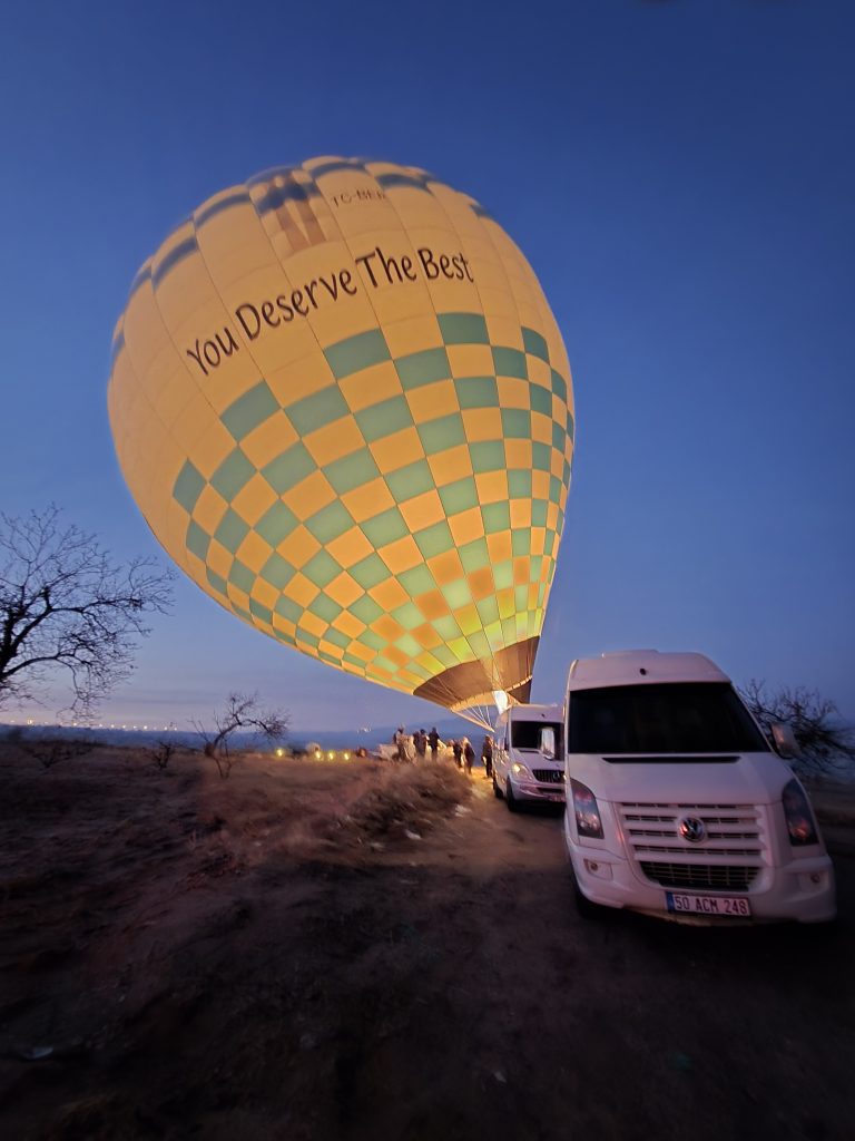 A large yellow and green hot air balloon being inflated at dawn in Cappadocia, with the phrase “You Deserve The Best” printed on it. Two white vans and several people are gathered nearby on a dirt clearing under a deep blue sky.