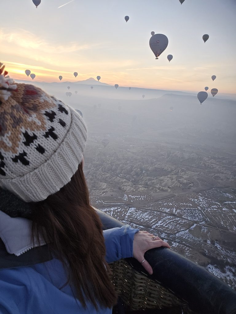 Me looking out from a hot air balloon basket at sunrise, surrounded by other balloons drifting across a misty Cappadocian valley. The warm glow highlights the unique volcanic formations below.