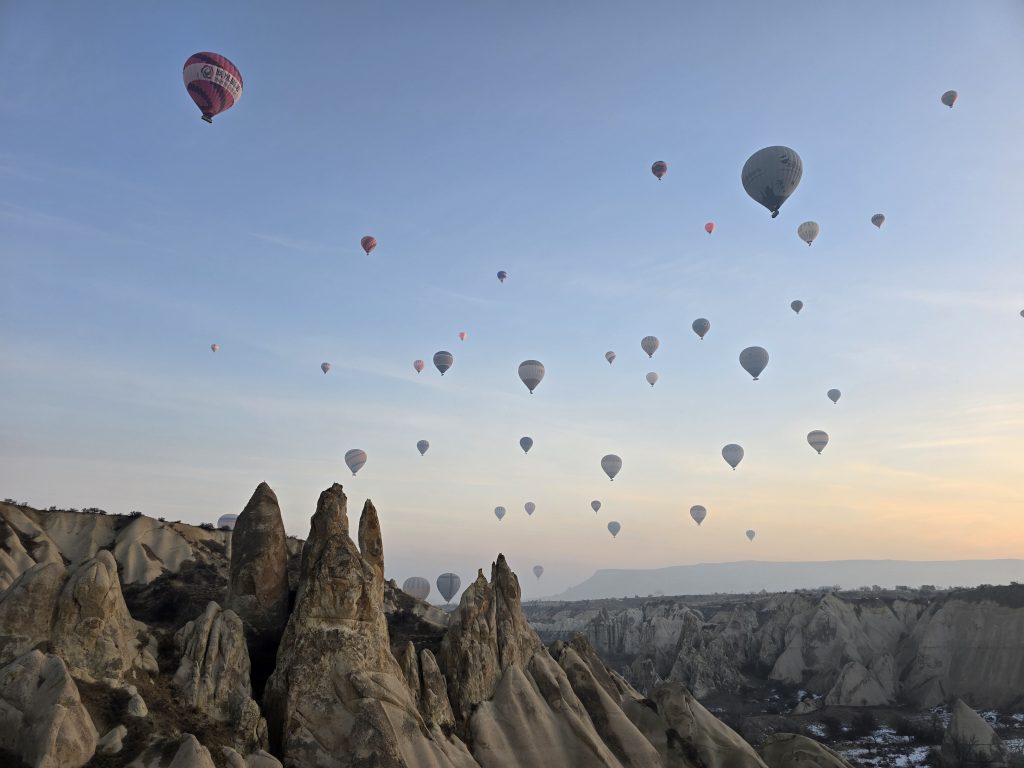 Cluster of hot air balloons rising into the morning sky over Cappadocia’s jagged rock formations. The stone spires in the foreground contrast with the soft, pastel sky.