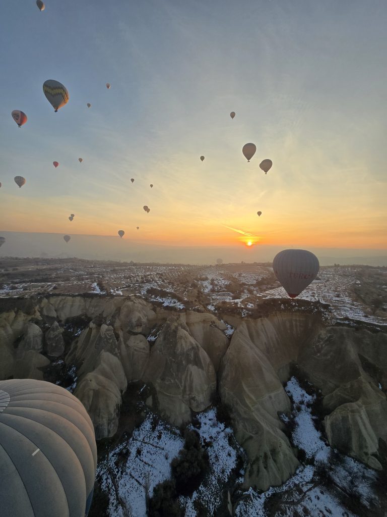 Hot air balloons soaring above the snow-dusted fairy chimneys of Cappadocia at sunrise. The golden morning light casts a warm glow over the surreal landscape, highlighting the region’s famous rock formations and the magical atmosphere of hot air balloon rides in Cappadocia.