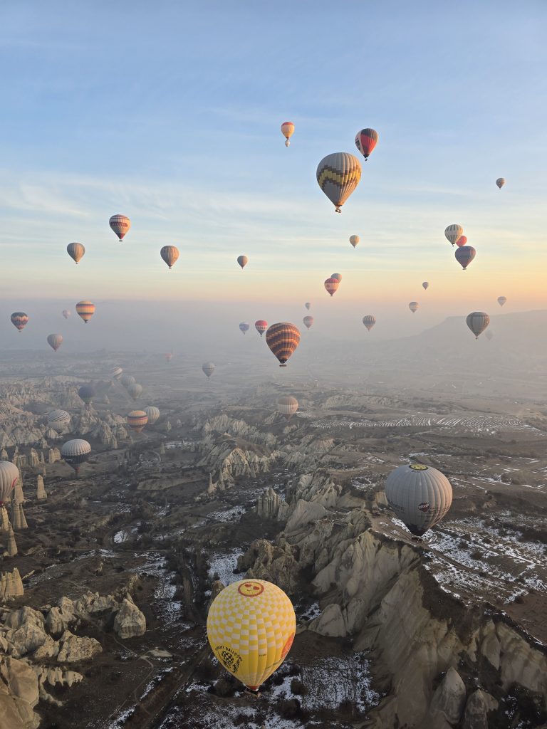Hot air balloons fill the sky above Cappadocia's snow-dusted valleys and rugged landscape during a sunrise flight. The variety of balloon colors and patterns adds to the magical view.