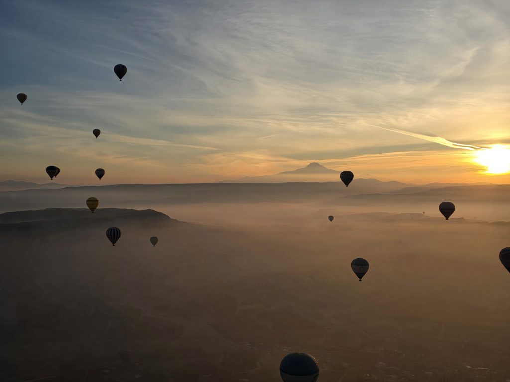 Silhouettes of hot air balloons float through the golden morning mist as the sun rises over the mountains in Cappadocia. Mount Erciyes appears faintly in the distance, adding depth to the scene.