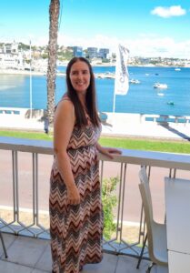 Me standing on a balcony at Baía do Peixe in Cascais, smiling in a chevron-patterned maxi dress with shades of rust, black, and white. Behind me, boats float on the blue waters of the bay with the Cascais skyline and waterfront in the background.