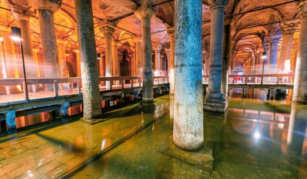 Wide view of the Basilica Cistern showing rows of ancient marble columns reflected in shallow water, with arched ceilings and moody orange lighting creating a dramatic atmosphere. Elevated walkways allow visitors to explore the historic underground reservoir.