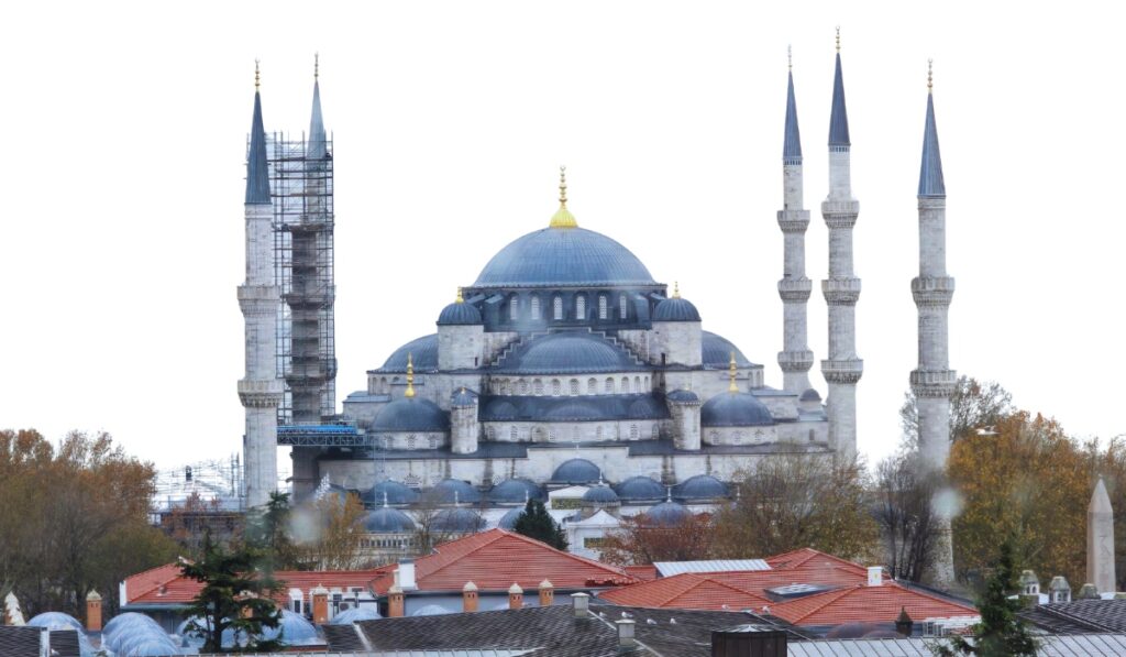 Daytime view of the Blue Mosque in Istanbul with all six minarets visible, one of which is under scaffolding. The central dome and cascading smaller domes are framed by autumn trees and red-roofed buildings in the foreground against an overexposed white sky.